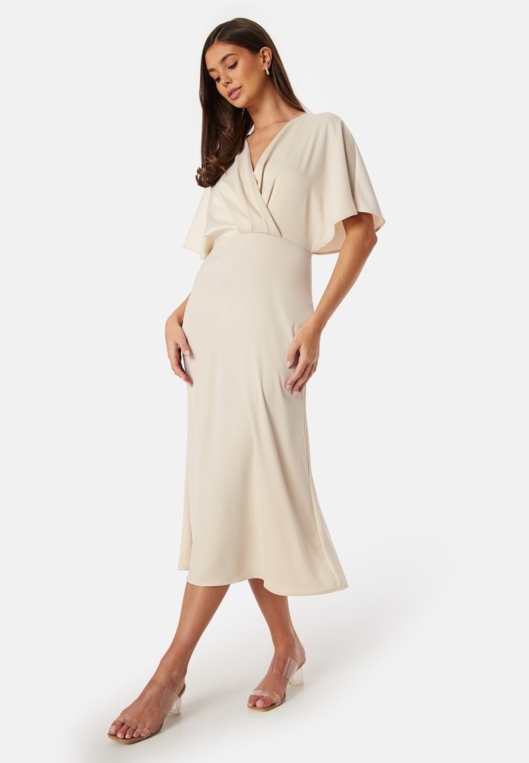 Beige knälång klänning med en omslagningsdesign, korta, svävande ärmar och mjuk textur; parat med klara sandaler med blockklack.