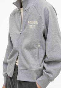 Grau melierte Reißverschlussjacke mit gerippten Bündchen und Kragen; mit hellbeigem "ROUGE EDIT"-Stickereimotiv. Verfügt über eine Seitentasche und eine weiche Textur.