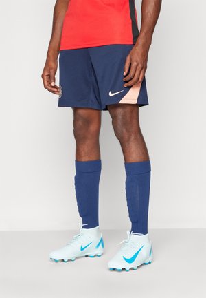 Marineblauwe sportshorts met lichtperzikkleurige accenten en een logo, gecombineerd met blauwe sokken en lichtblauwe voetbalschoenen met een blauwe swoosh.