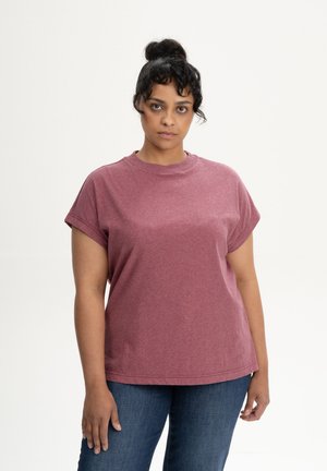 Frau mit dunkel gebundenen Haaren, die ein kurzärmeliges mauvefarbenes T-Shirt und blaue Jeans trägt und vor einem schlichten hellen Hintergrund steht.