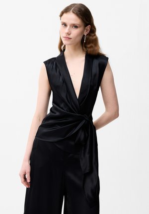 Giovane donna con capelli mossi che indossa un top nero in raso senza maniche incrociato e pantaloni a gamba larga, in piedi davanti a uno sfondo bianco uniforme.