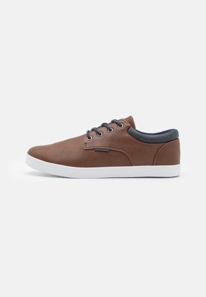 Zapatilla de cuero marrón con un perfil bajo, suela de goma blanca, punta redonda, detalles azules en el collar y seis pares de ojetes para los cordones.