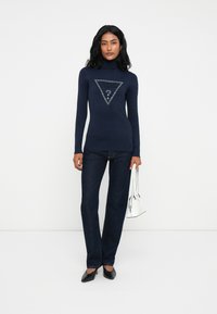 Mujer vistiendo un suéter de cuello alto en navy con el logo "GUESS", jeans oscuros y tacones negros, sosteniendo un bolso blanco frente a un fondo blanco liso.