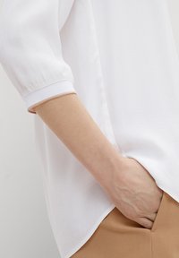 Blusa bianca con maniche a trequarti, caratterizzata da un tessuto a trama e polsini a coste in contrasto. Abbinata a pantaloni beige, con una mano in tasca.