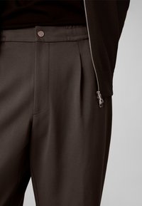 Gros plan sur un pantalon marron foncé sur mesure avec des plis latéraux, une fermeture à bouton argenté, et une partie d’une veste noire zippée.