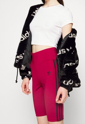 Kobieta ubrana w biały krótki top, czarną puszystą kurtkę z białym nadrukiem oraz czerwone, wysokowycięte spodenki kolarskie Adidas z czarnymi paskami.