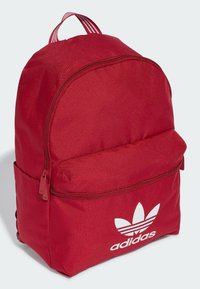 Mochila roja de Adidas hecha de tela texturizada. Presenta una parte superior redondeada, compartimento principal con cremallera y bolsillo frontal con logo blanco. Asa a rayas.