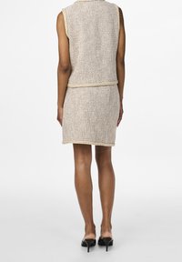 Béžový pletený dvoudílný outfit s bezrukávovým topem a přiléhavou sukní, doplněný kontrastním lemem podél okrajů.