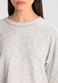Sudadera gris clara hecha de tejido texturizado, con un cuello redondo y mangas raglán, que presenta sutiles variaciones en color y patrón.