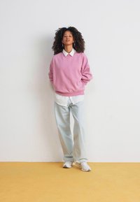 Rosa Baumwollpullover mit V-Ausschnitt, über einem weißen Hemd mit Kragen getragen; hellblaue weite Hose; weiße Turnschuhe; auf gelbem Boden stehend.