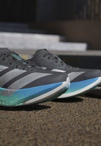 Coppia di scarpe da corsa grigio scuro e verde acqua con strisce bianche posizionate su una superficie esterna con struttura vicino a dei gradini.