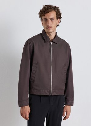 Man with curly hair wearing a dark brown zip-up jacket and black pants, standing against a plain light gray wall.