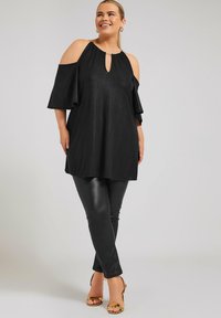 Top nero con spalle scoperte e scollatura a chiave, maniche svasate e finitura testurizzata, abbinato a pantaloni neri aderenti e lucidi e tacchi dorati.