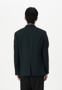 Dunkelgrüner maßgeschneiderter Blazer mit glatter Textur, Reverskragen und einer Schlitze hinten, kombiniert mit dunklen Hosen.