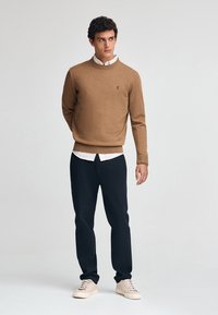 Brauner Strickpullover mit rundem Ausschnitt, getragen über einem weißen Hemd mit Kragen, kombiniert mit dunklen Hosen und beigen Schuhen, vor einem neutralen Hintergrund stehend.