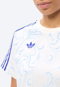 Inimene, kes kannab valget Adidas t-särki sinise keerutusega mustriga ja kolme sinise triibuga õlal, pildistatud lõualuu ja rinna tasandil.