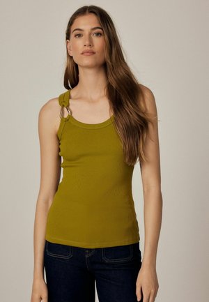 Femme aux longs cheveux bruns portant un haut sans manches vert olive avec des détails en anneaux métalliques et un jean bleu foncé, debout devant un fond uni.