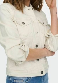 Beige denimjacka med krage, knäppning framtill, två bröstfickor och uppvikta ärmar. Har metallknappdetaljer och en strukturerad passform.