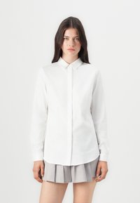 Chemise blanche à manches longues avec un front boutonné et un col pointu. Texture lisse avec un ourlet arrondi. Associée à une jupe grise plissée.