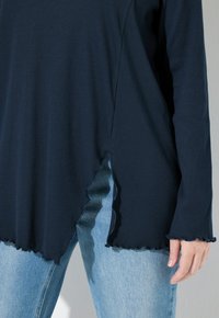 Top a maniche lunghe a coste blu navy con orlo scallop e spacchi laterali, abbinato a jeans azzurri. La visuale ravvicinata evidenzia la trama e le cuciture.