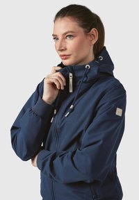 Marinblå zip-up jacka med huva, elastiska ärmslut, dragsnören och en logotyp på ärmen, tillverkad av väderbeständigt material.