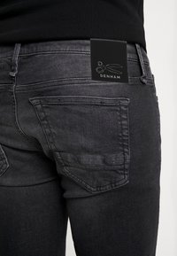 Primer plano de una persona que lleva puestos unos pantalones vaqueros de mezclilla negros desteñidos con un bolsillo trasero y una etiqueta de la marca Denham que muestra el logo de unas tijeras.