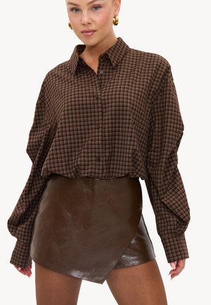Femme portant une chemise à carreaux marron à manches longues et une mini-jupe en cuir marron foncé avec des boucles d’oreilles anneaux dorés sur fond blanc.