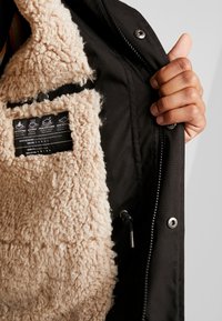 Chaqueta exterior negra con un forro aislante de color marrón claro y suave. Cuenta con una etiqueta de cuidado y un bolsillo con cremallera en el interior. Acentos de herrajes plateados.