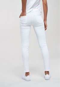 Persona in jeans skinny bianchi e sneakers bianchi, in piedi con una mano nella tasca posteriore contro uno sfondo chiaro e semplice.