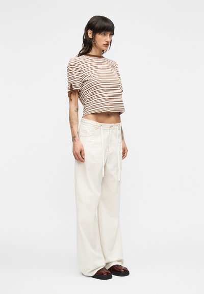 Γυναίκα με σκούρα μαλλιά φοράει κοντομάνικη ριγέ μπλούζα σε καφέ και λευκό, παντελόνι ευρύ και ψηλόμεσο σε λευκό χρώμα, και σκούρα καφέ παπούτσια, στέκεται μπροστά σε λευκό φόντο.