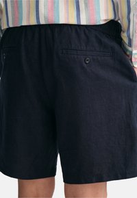 Shorts en lin bleu marine avec une taille élastique, dotés d'une poche arrière unique avec un bouton. Le tissu a une texture douce et un éclat minimal.