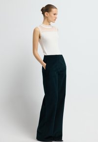 Krémový žebrovaný bezramínkový top s průhlednými volánkovými detaily kolem výstřihu, spárovaný s tmavozelenými kalhotami s širokými nohavicemi a předními kapsami.