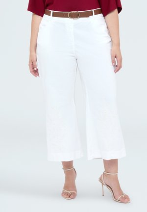 Personne portant un pantalon blanc large et court avec une broderie discrète, une ceinture marron, un haut rouge et des sandales beiges à talons hauts avec des brides.