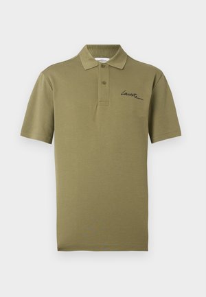 Мъж и жена, облечени с маслиненозелени поло тениски и бели долнища, стоящи един до друг пред светъл равен фон.