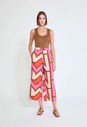 Mujer de pie con las manos en los bolsillos, vistiendo un top marrón sin mangas y pantalones de pierna ancha con un patrón geométrico llamativo en rosa, rojo y beige, y sandalias marrones.