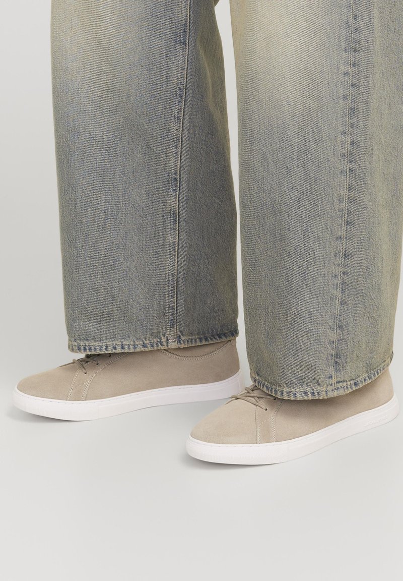 Adidași din suede bej cu tălpi din cauciuc alb, având șireturi și detalii de cusătură, purtați cu blugi din denim albastru deschis, cu croială largă.