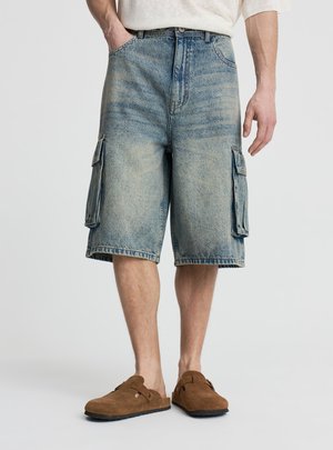 Mężczyzna w wyblakłych, niebieskich dżinsowych szortach typu cargo i brązowych zamszowych butach wsuwanych, stojący na tle jasnej, jednolitej ściany.