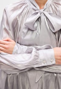 Silvery blouse with a bow tie collar, flowing fabric, gathered details, and buttoned cuffs adorned with circular embellishments.