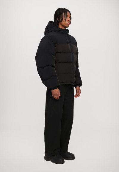 Sort pufferjakke med hætte, der har en quiltet tekstur og en frontlynlås. Bæret med løse sorte bukser og slip-on sko.
