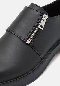 Scarpa in pelle nera senza lacci con chiusura a zip, superficie liscia, cuciture contrastanti e suola in gomma con un leggero tacco.