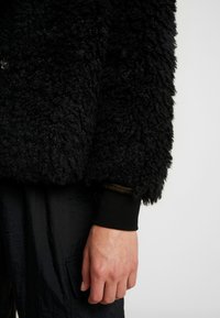 Veste noire texturée avec un matériau duveteux. Comprend des poignets noirs côtelés et des coutures visibles. La main repose sur un tissu sombre et brillant.