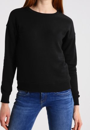 Person, die einen schlichten schwarzen Langarmpullover und blaue Jeans trägt, vor einem neutralen hellen Hintergrund stehend.