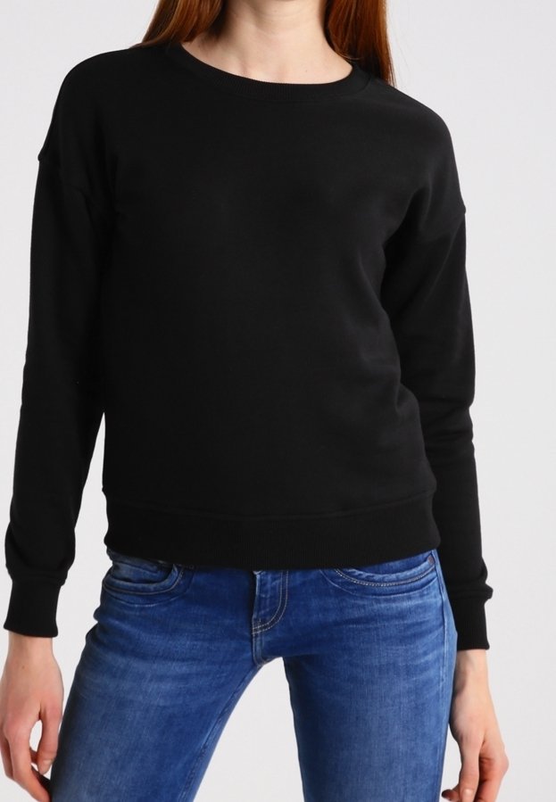 Personne portant un pull noir uni à manches longues et un jean bleu, debout devant un fond clair neutre.