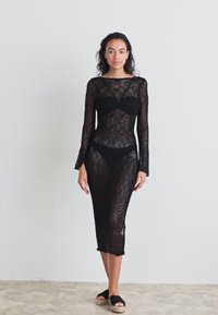 Vestito nero a maglia con maniche lunghe, silhouette aderente, lunghezza fino al polpaccio, trama a lavorazione aperta e bordi sfilacciati. Indossato sopra un bikini coordinato.