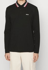 Svart långärmad pikétröja med en randig krage i röd, vit och svart, med en knäppning framtill och en logotyp på bröstet.