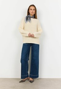 Maglione di crema spesso a maglia con maniche oversize, abbinato a jeans a gamba larga blu scuro e tacchi a righe bianche e nere, accessoriato con una sciarpa a fantasia.