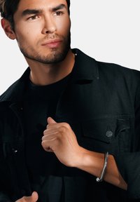 Svart denimjacka över en svart t-shirt, modell bär en silverarmband, fokuserad uttryck, neutral bakgrund, som betonar klädernas texturer.