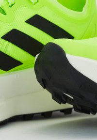 Zöld futócipők fekete csíkokkal, hálós felsőrésszel, texturált anyaggal és vastag fehér középtalppal. Gumitalp, amely tapadásmintákkal rendelkezik.