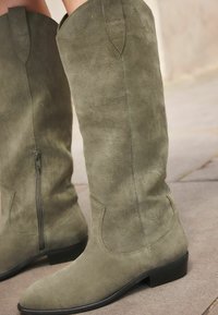 Botas de ante verde oliva hasta la rodilla con costuras decorativas, cremallera lateral y tacón bajo. Textura suave con una silueta ajustada.