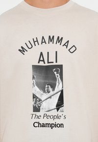 Beige T-krekls ar melnbaltu fotogrāfiju, kurā redzams Muhameds Ali ar paceltām dūrēm, un uzraksts "Muhameds Ali, Tautas čempions."